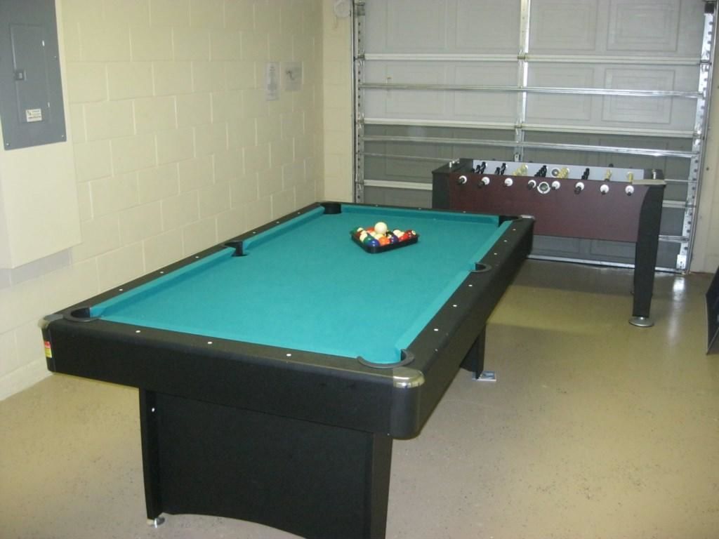 Ett biljardbord med en grön filtduk och ett fotbollsspelbord i ett rum med beige väggar, och en garageport syns i bakgrunden.