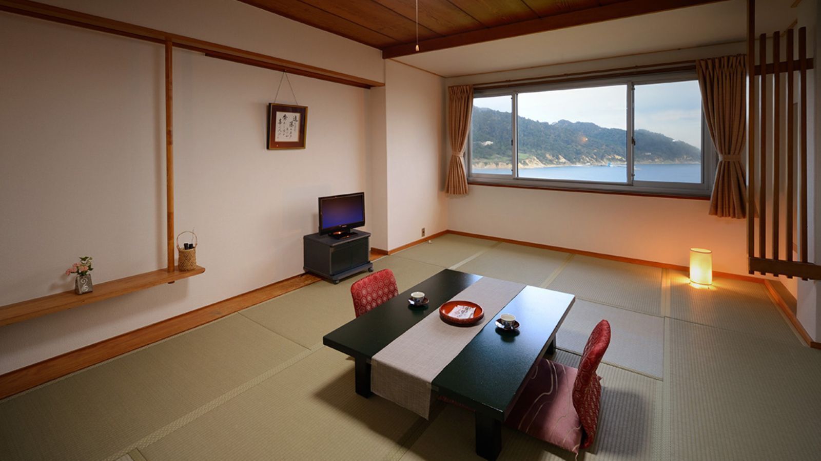 A traditional Japanese room with tatami flooring, a low table with a tea set, a television, and a view of the sea.