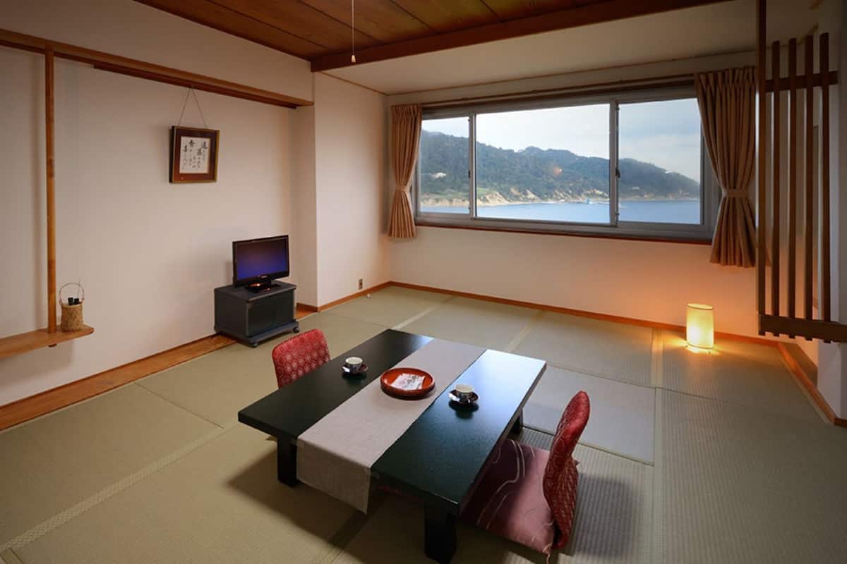 A traditional Japanese room with tatami flooring, a low table with a tea set, a television, and a view of the sea.