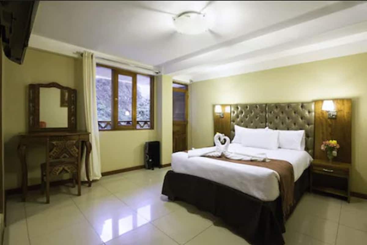 Habitación de hotel con una cama grande, una mesita de noche de madera, un espejo y una ventana con cortinas.