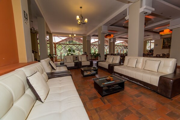 A spacious lounge area with leather sofas and armchairs, a coffee table, and large windows offering views of greenery.