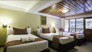 Habitación de hotel con tres camas, techo de madera y un amplio ventanal con vistas a árboles.