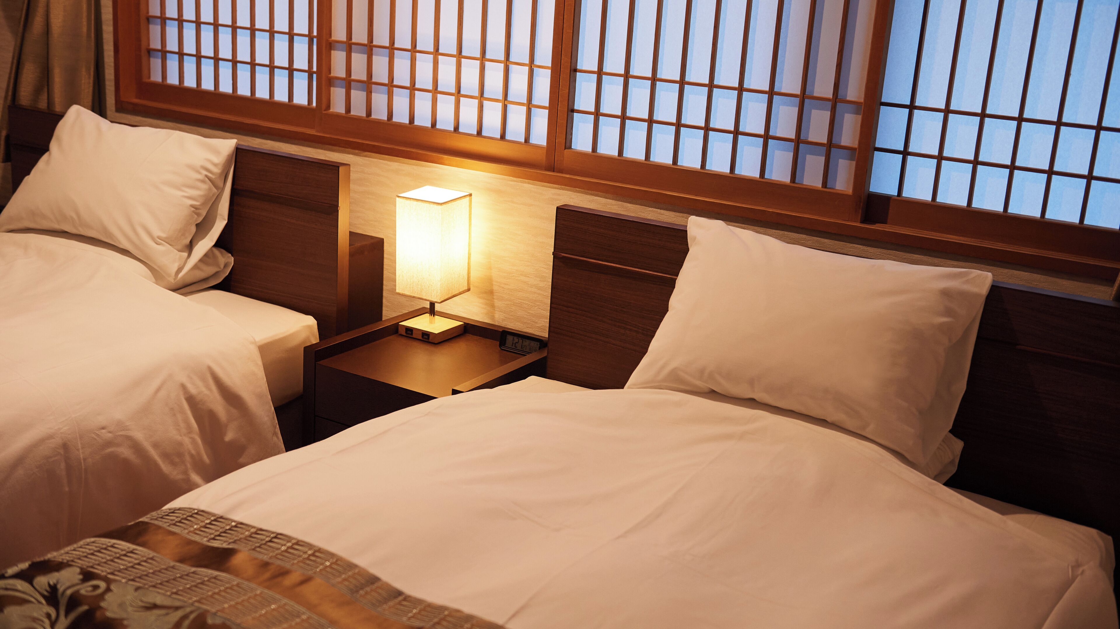 Una habitación de hotel con dos camas, una mesita de noche con lámpara y una ventana con persianas de madera.