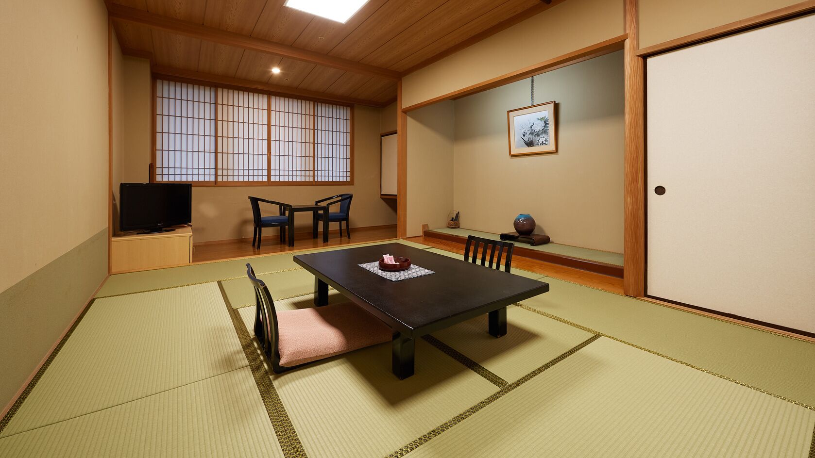 Una habitación japonesa tradicional con suelo de tatami, una mesa baja, sillas, un televisor y una puerta corredera.