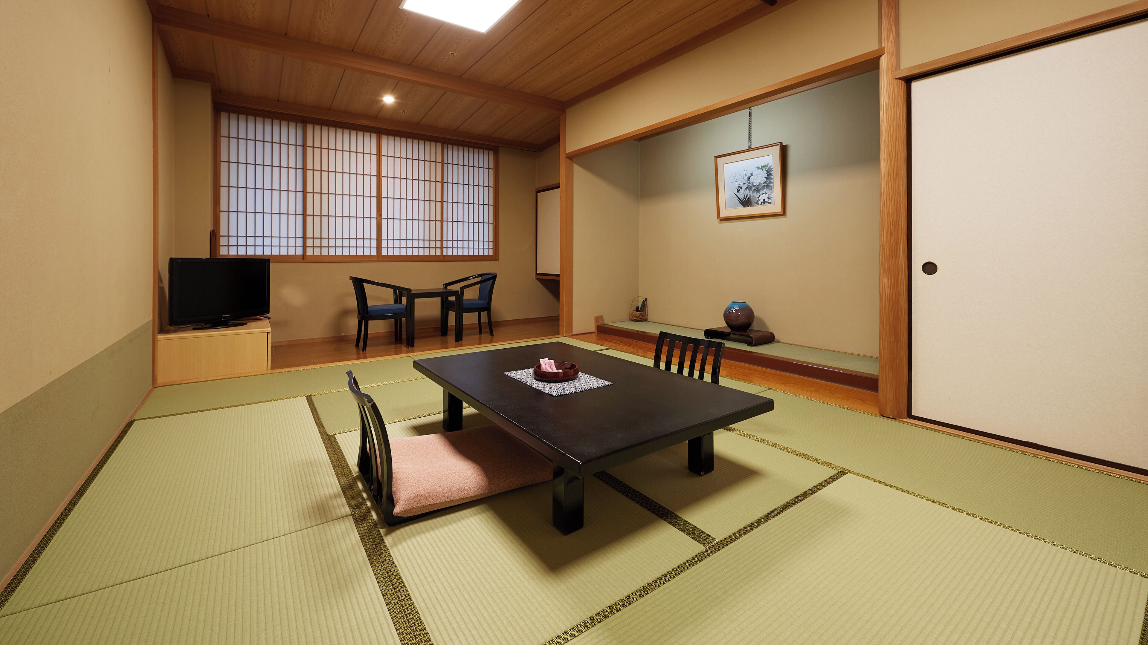Una habitación japonesa tradicional con suelo de tatami, una mesa baja, sillas, un televisor y un cuadro en la pared.