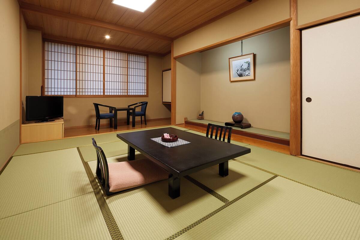 Une pièce de style japonais traditionnel, avec un sol recouvert de tatamis, une table basse, des chaises, un téléviseur et un tableau encadré accroché au mur.