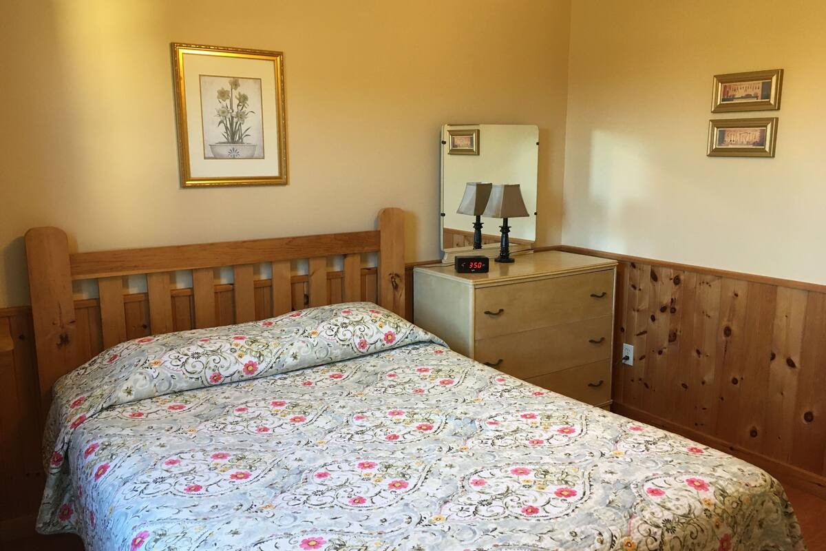 A bedroom with a wooden headboard, a bed with floral bedding, a dresser with a mirror, and two framed pictures on the wall.