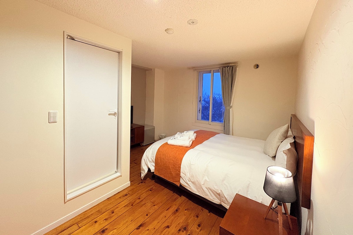 A bedroom with a wooden floor, a bed with white bedding, a bedside table with a lamp, a window with curtains, and a door.
