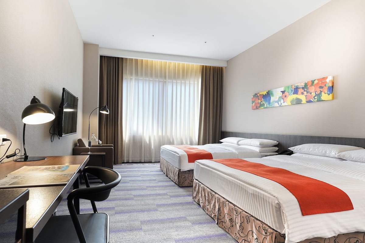A hotel room with two beds, a desk with a lamp, a TV, and a large window with curtains.