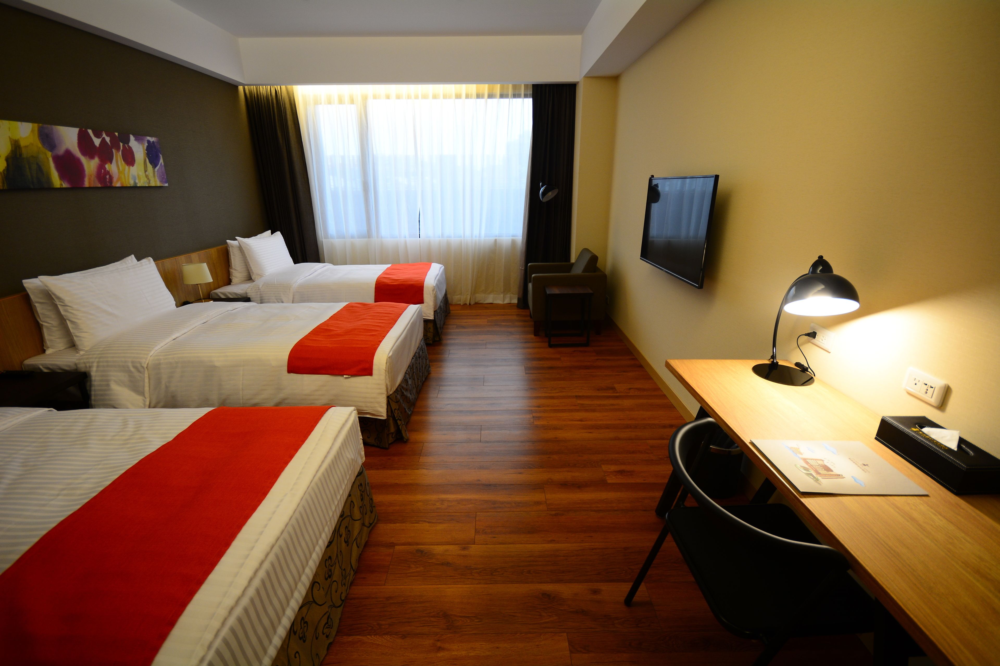 Habitación de hotel con tres camas, un escritorio, una silla, un televisor y una lámpara.