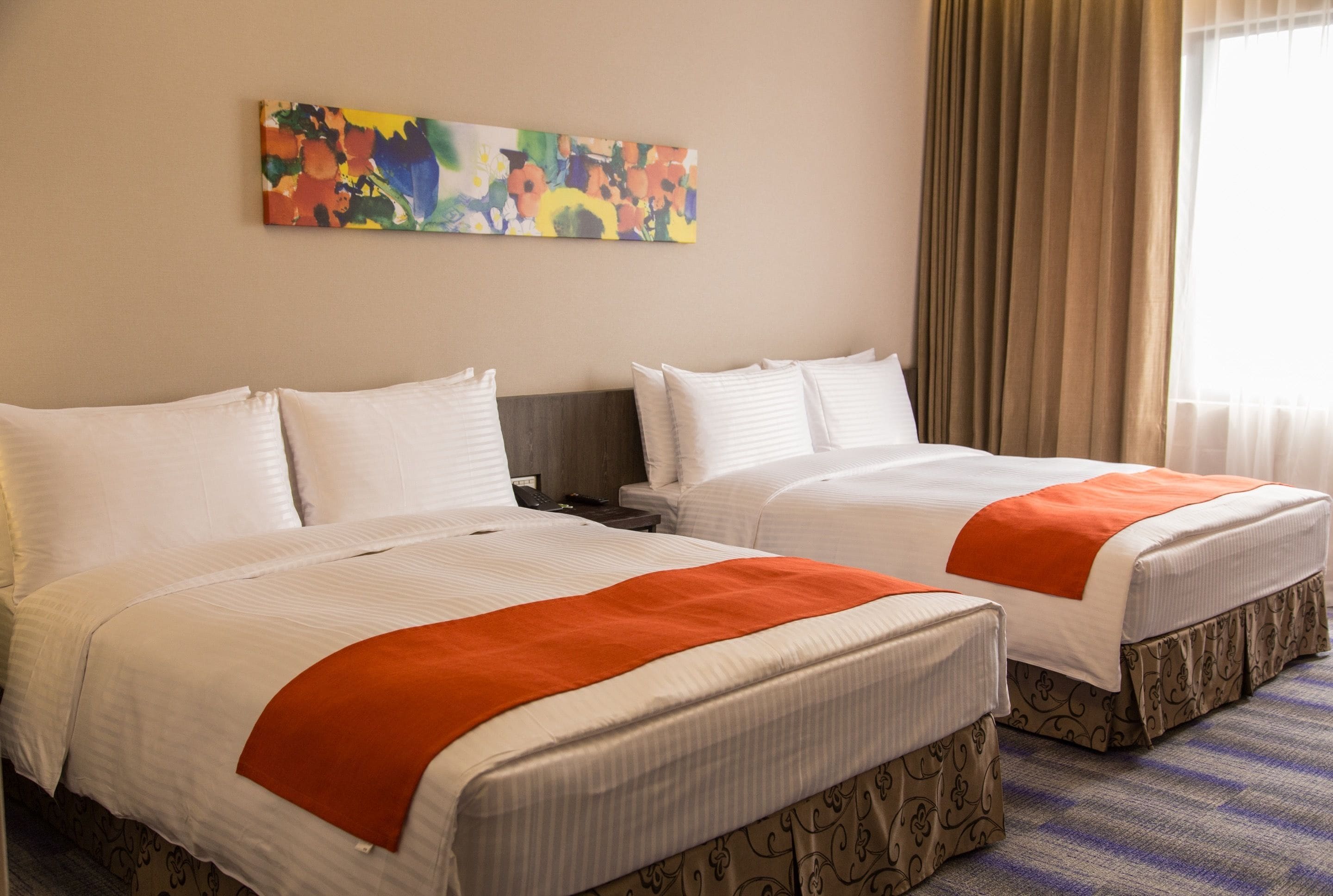A hotel room with two beds, a nightstand, a painting on the wall, and a window with curtains.