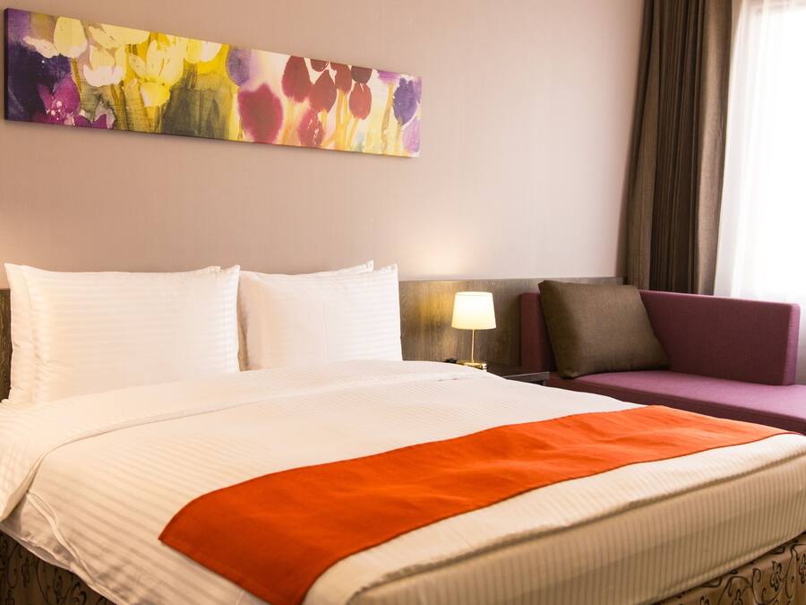A hotel room with a large bed, a purple armchair, a nightstand with a lamp, and a colorful abstract painting on the wall.