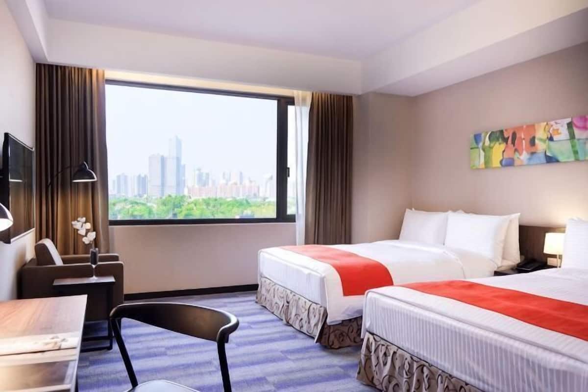 A hotel room with two beds, a desk, a chair, a TV, and a large window with a city view.