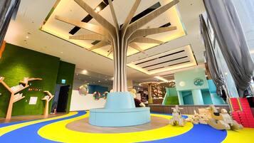 Un espacio interior moderno con una estructura arbórea central, zona de juegos colorida y diferentes disposiciones de asientos.