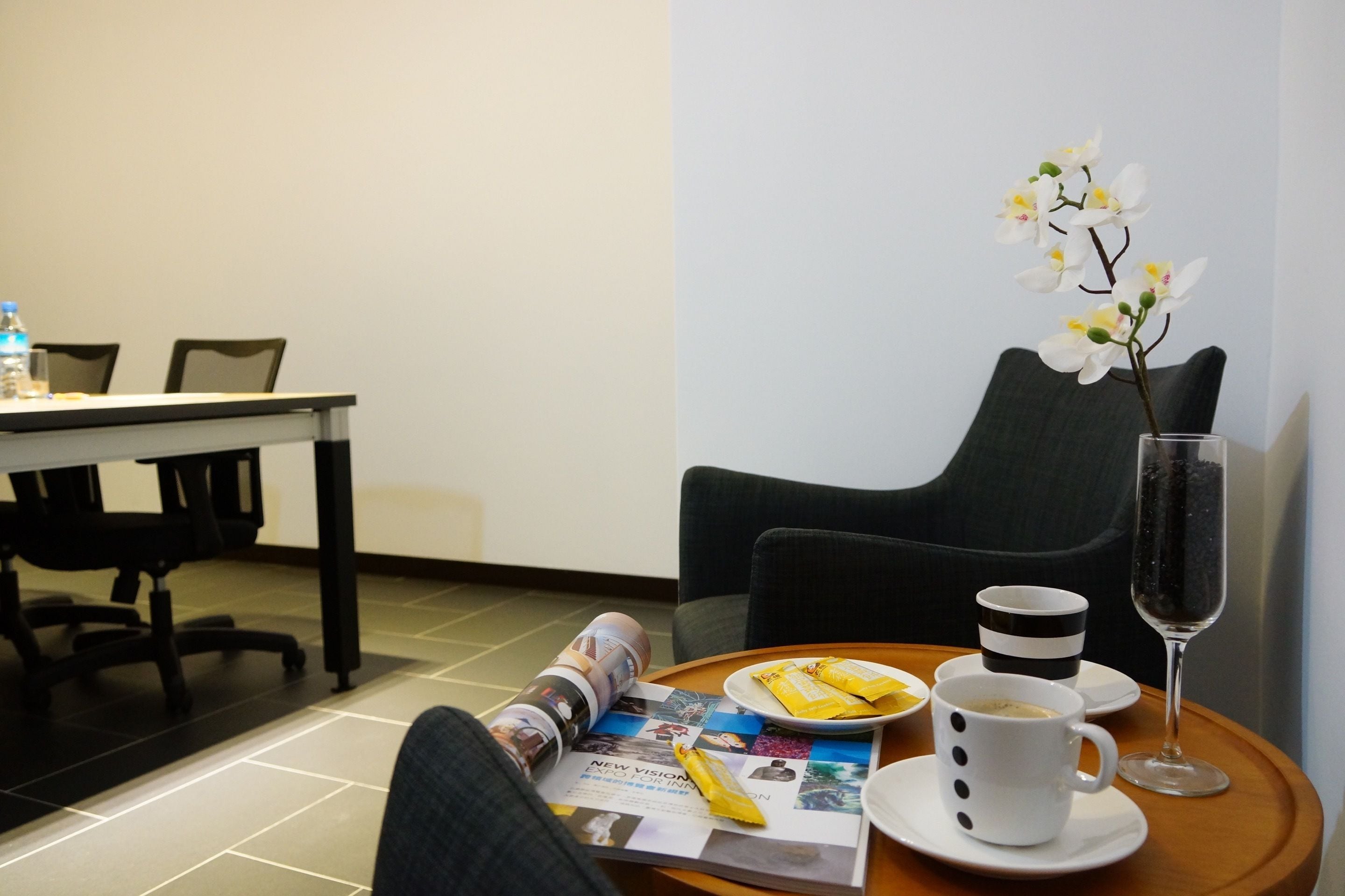 A hotel room with a dining table, a magazine, a plate with food, a cup of coffee, a glass of water, and a vase with white flowers.
