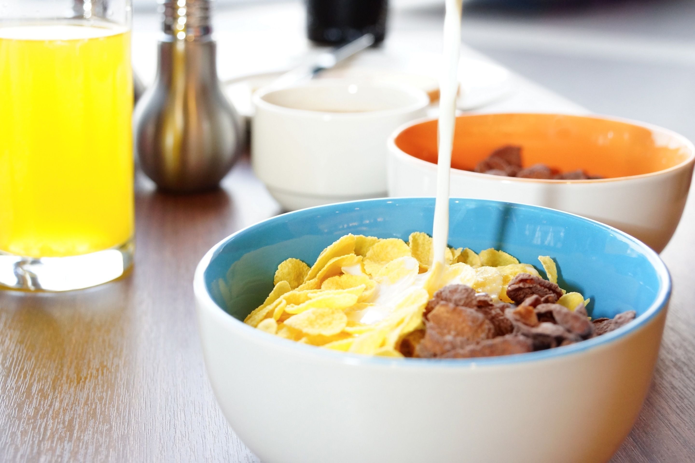 Een ontbijttafel met kommen vol cornflakes en melk, een glas sinaasappelsap en zout- en peperstrooiers op een houten tafel.