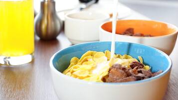 Een ontbijttafel met kommen vol cornflakes en melk, een glas sinaasappelsap en zout- en peperstrooiers op een houten tafel.