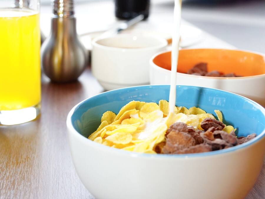 Een ontbijttafel met kommen vol cornflakes en melk, een glas sinaasappelsap en zout- en peperstrooiers op een houten tafel.