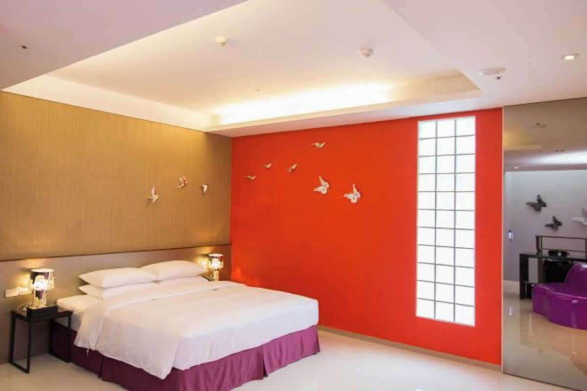 A modern hotel room with a large bed, a red accent wall, and a large window.