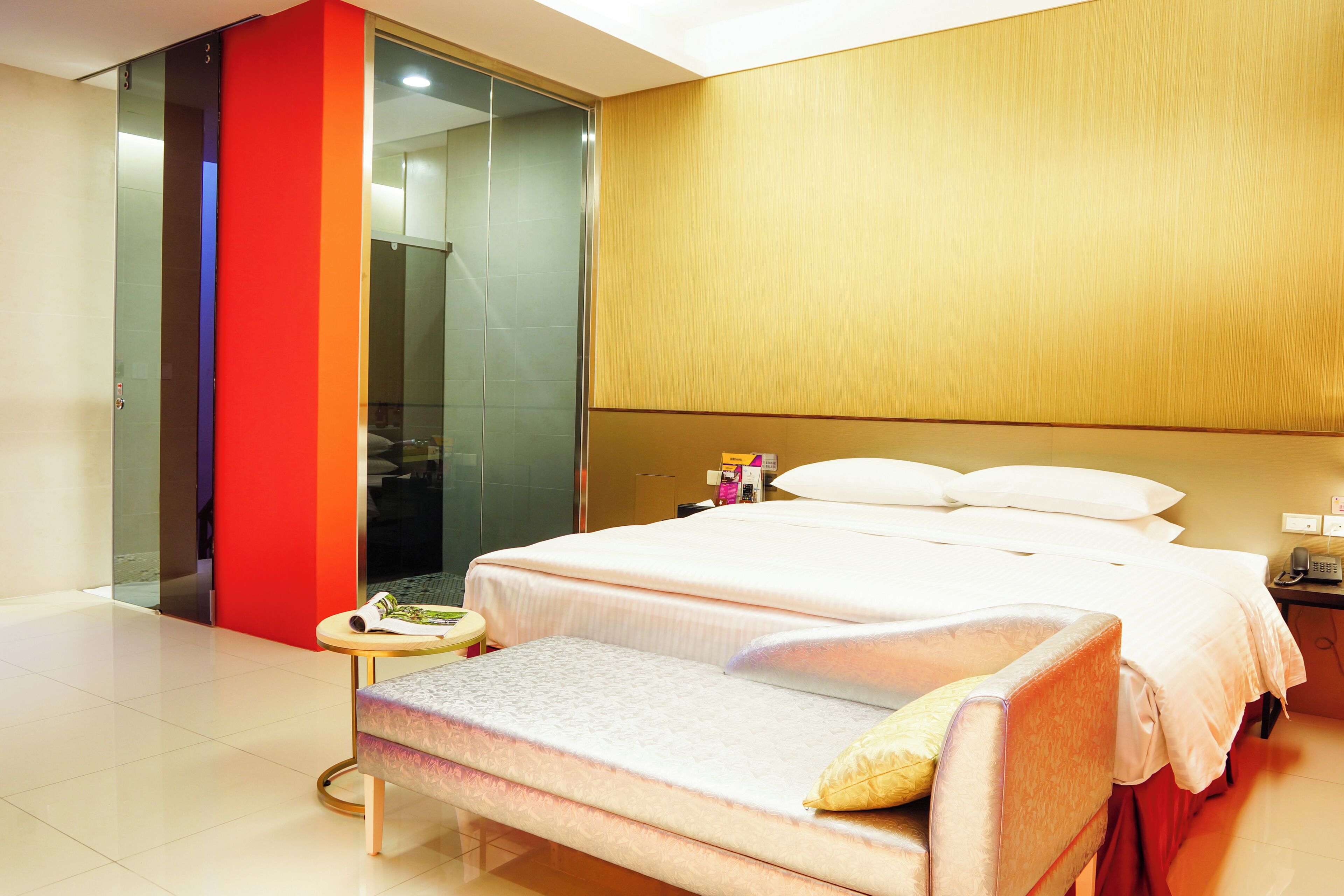 A modern hotel room with a large bed, a bedside table, a small stool, and a glass shower area.