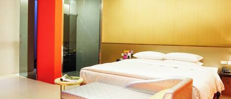 A modern hotel room with a large bed, a bedside table, a small stool, and a glass shower area.