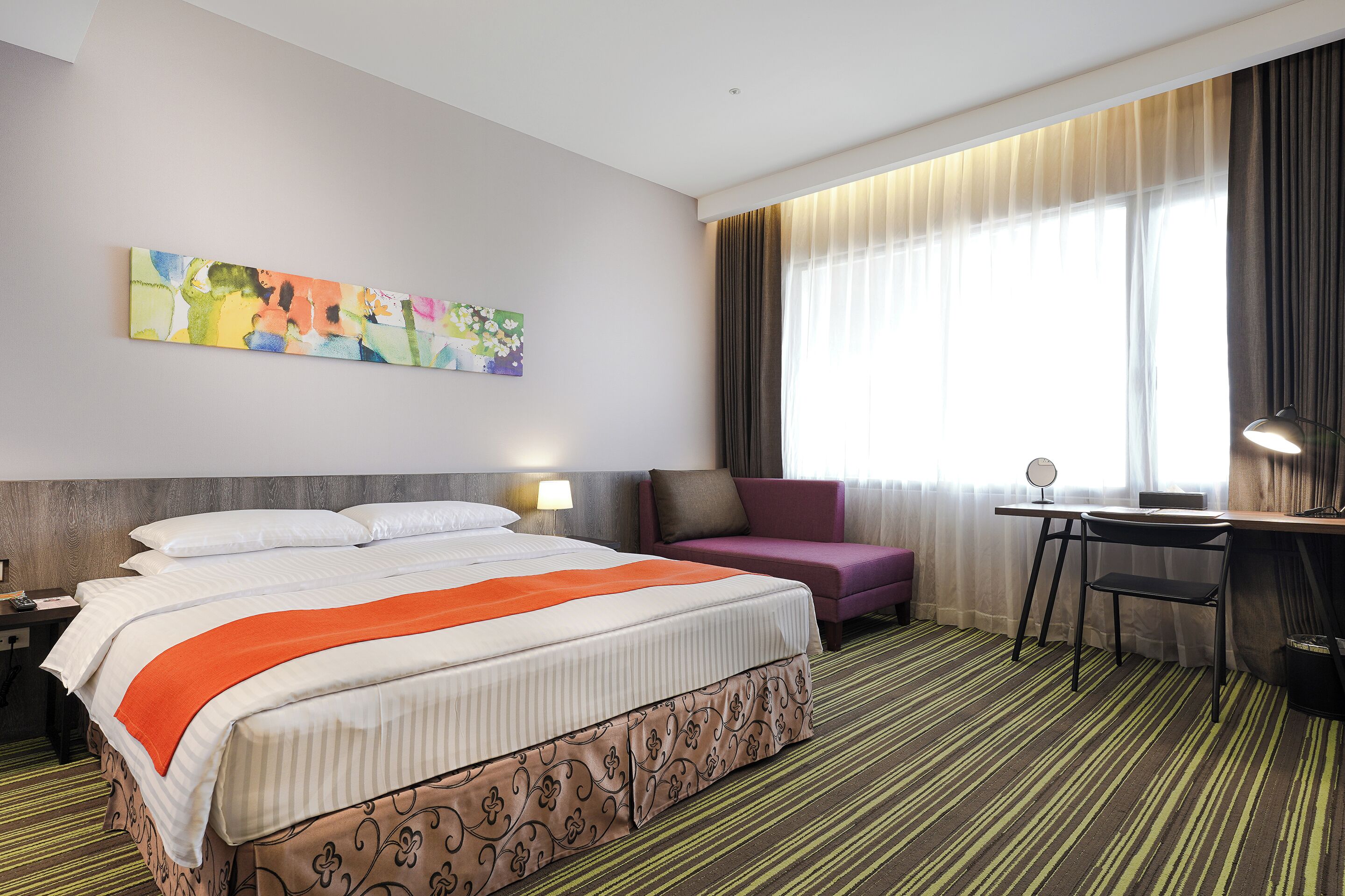 A hotel room with a bed, a desk, a chair, a sofa, and a large window with curtains.