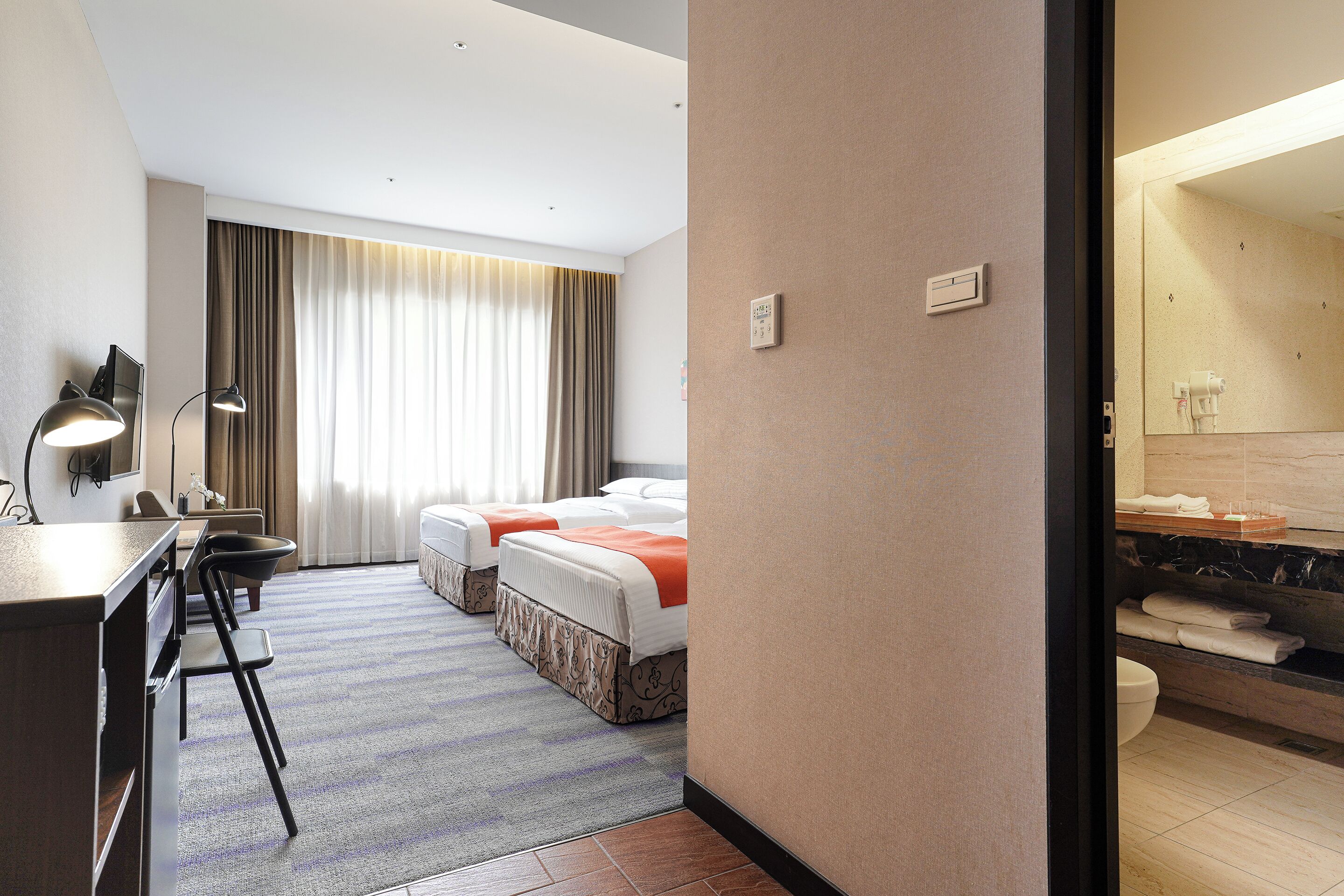 A hotel room with two beds, a desk, a chair, a desk lamp, a large window with curtains, and a bathroom with a mirror and towel rack.