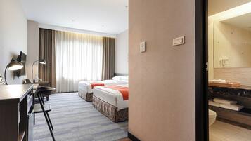 A hotel room with two beds, a desk, a chair, a desk lamp, a large window with curtains, and a bathroom with a mirror and towel rack.