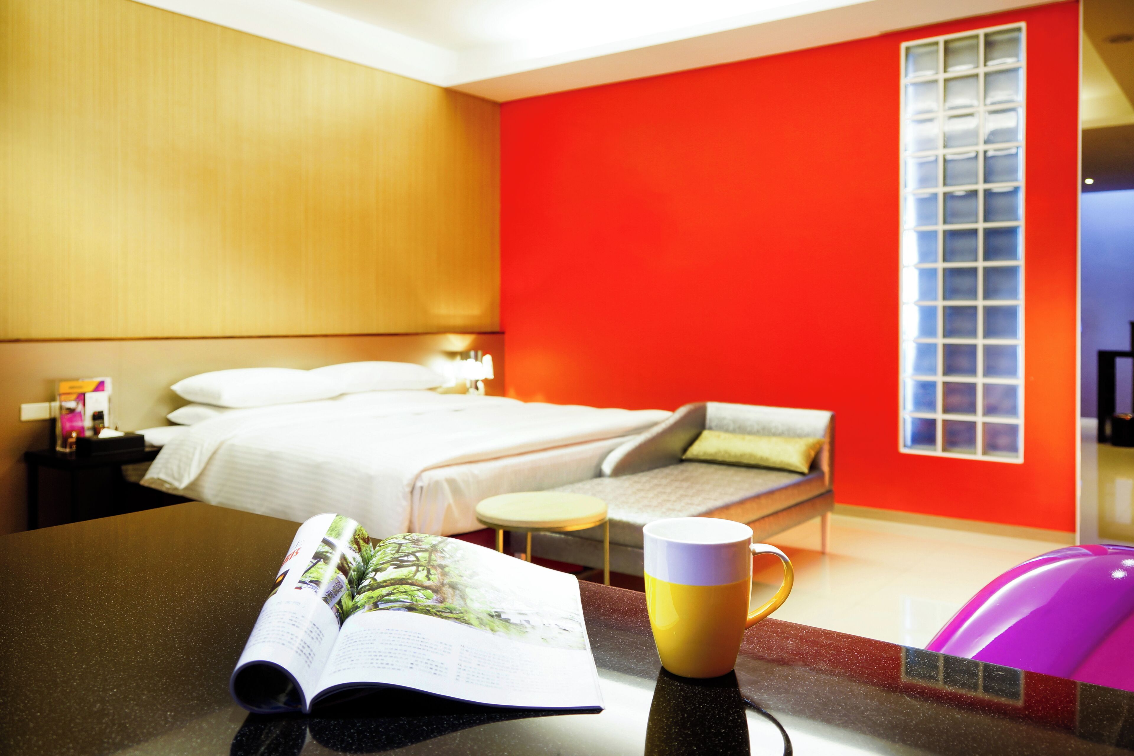 Una habitación de hotel con una cama, una mesita pequeña con una revista y una taza, una mesita de noche y una ventana con persianas.