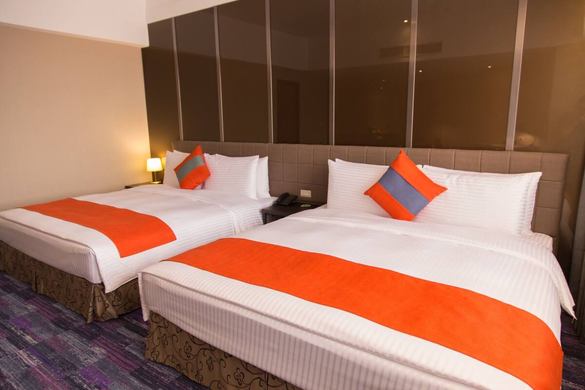 Two double beds with white and orange bedding, a nightstand, and a large window with blinds.