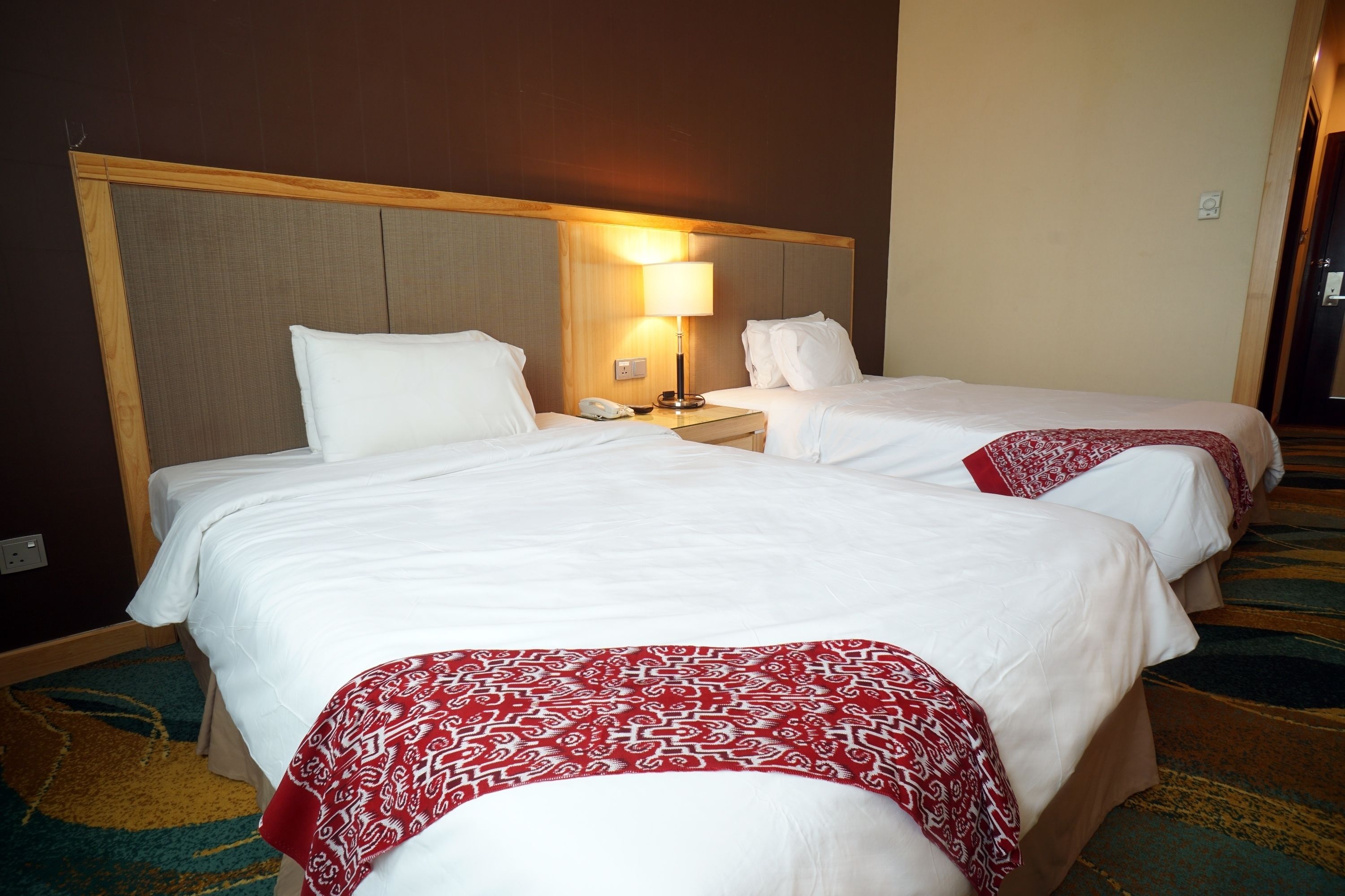 A hotel room with two beds, a nightstand with a lamp, and a carpeted floor.