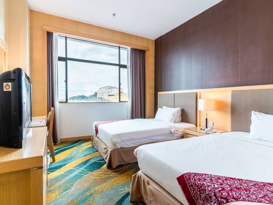 A hotel room with two beds, a desk with a television, a window with a view, and a patterned carpet.