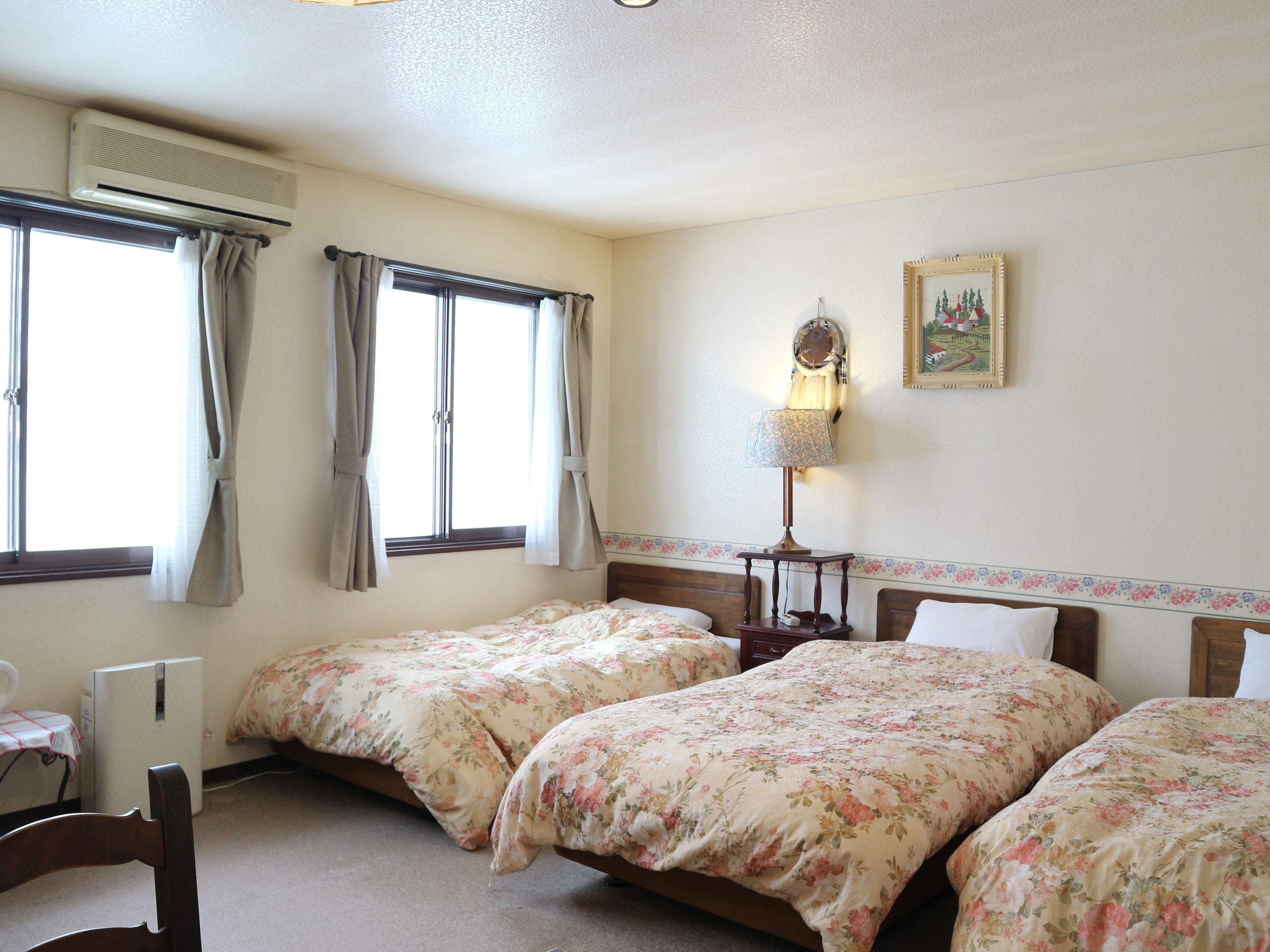 A room with two single beds, a desk, a chair, an air conditioner, and a wall-mounted lamp.
