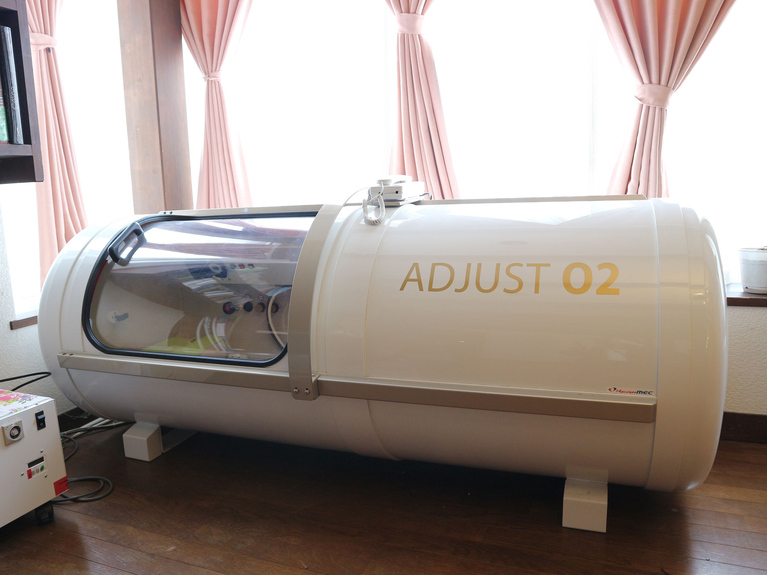 Un dispositivo médico etiquetado como "ADJUST 02" en una habitación con piso de madera y cortinas rosadas.