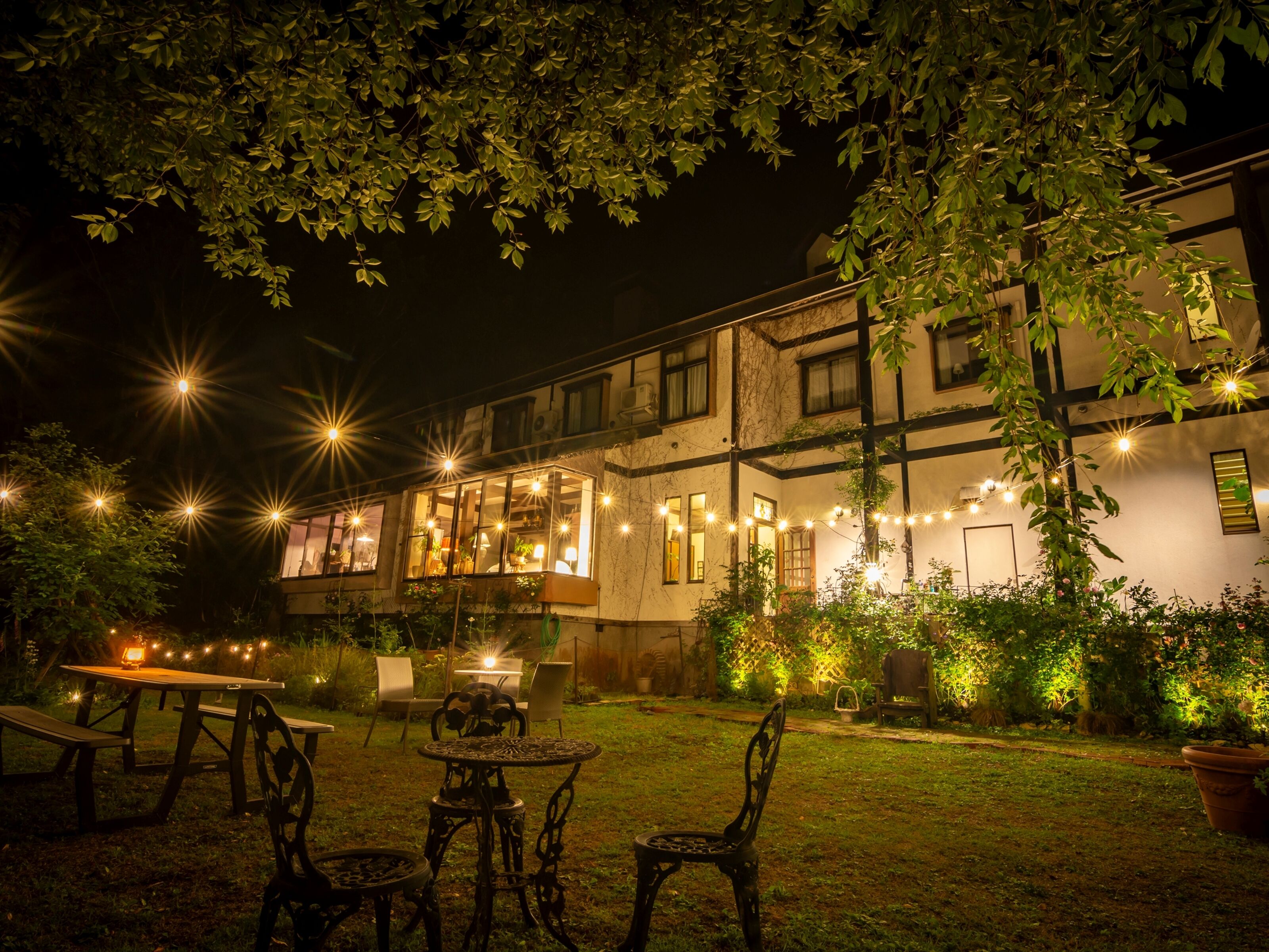 An outdoor seating area with tables and chairs, a building with multiple windows, and string lights.