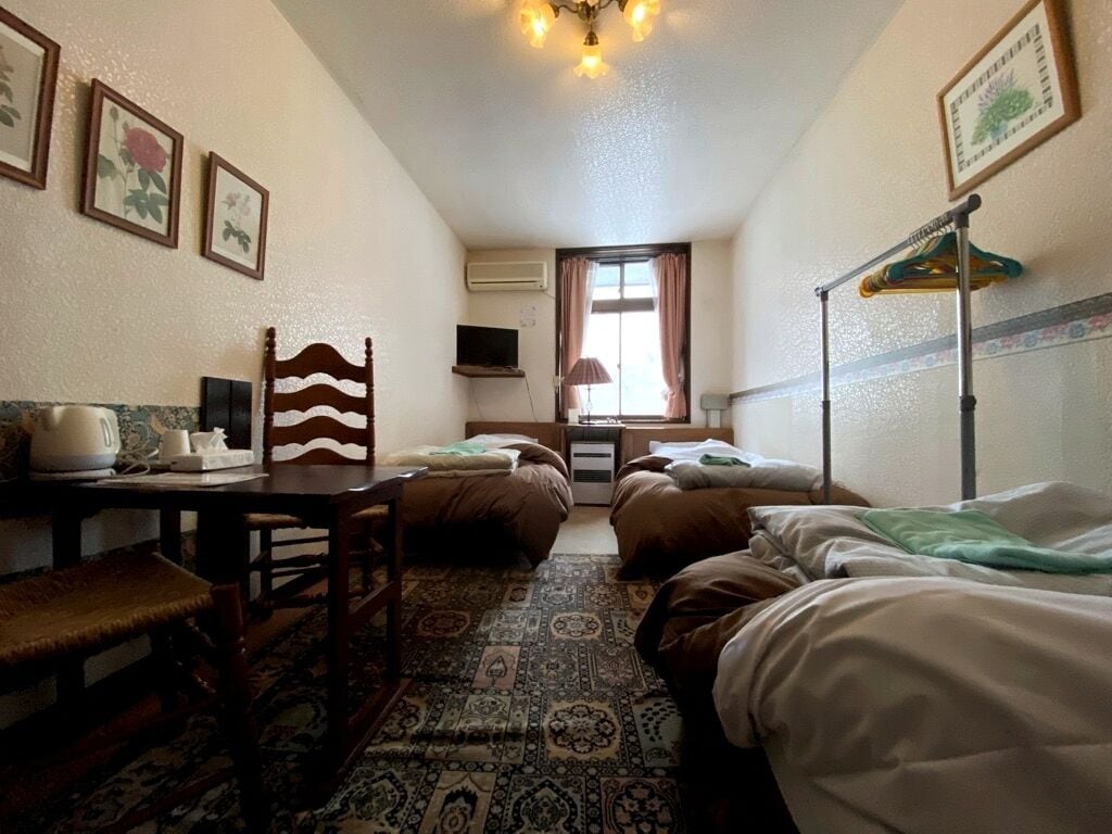 A small, cozy room with two single beds, a wooden chair, a table with a kettle, and a TV on the wall.