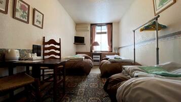 A small, cozy room with two single beds, a wooden chair, a table with a kettle, and a TV on the wall.