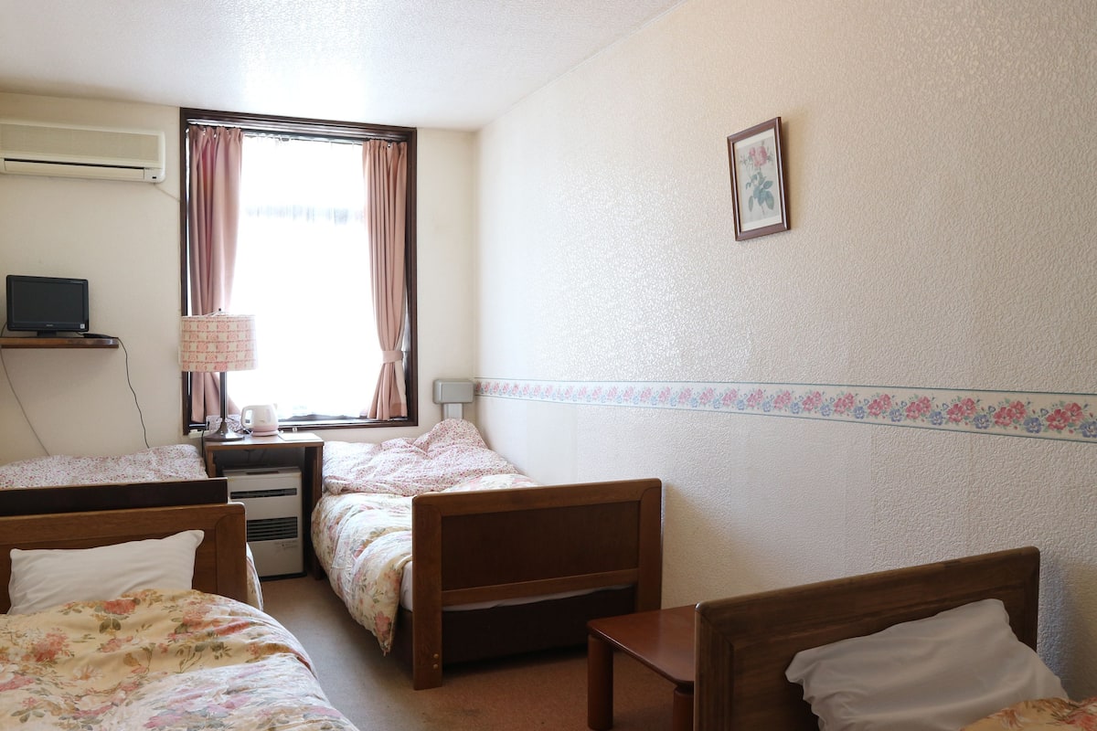 Habitación de hotel con dos camas individuales, un escritorio con lámpara, televisión y ventana con cortinas.