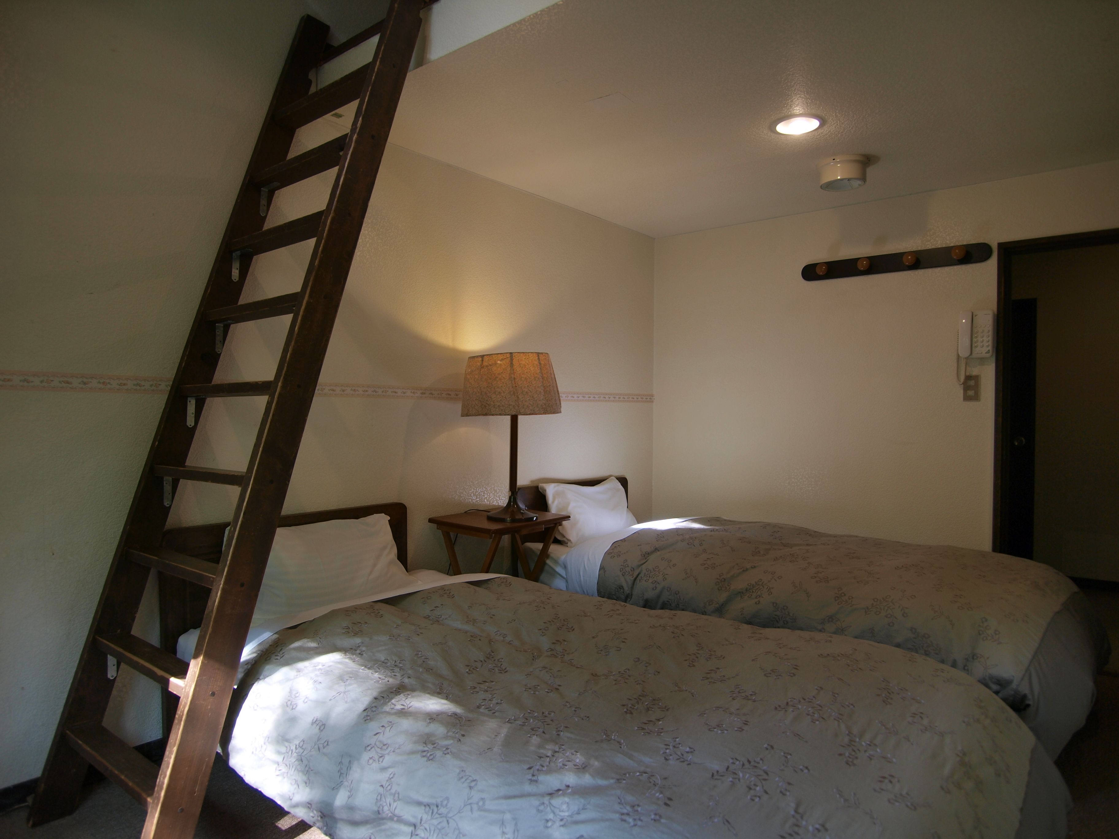 Een slaapkamer met twee bedden, een nachtkastje, een lamp, een telefoon en een ladder tegen de muur.