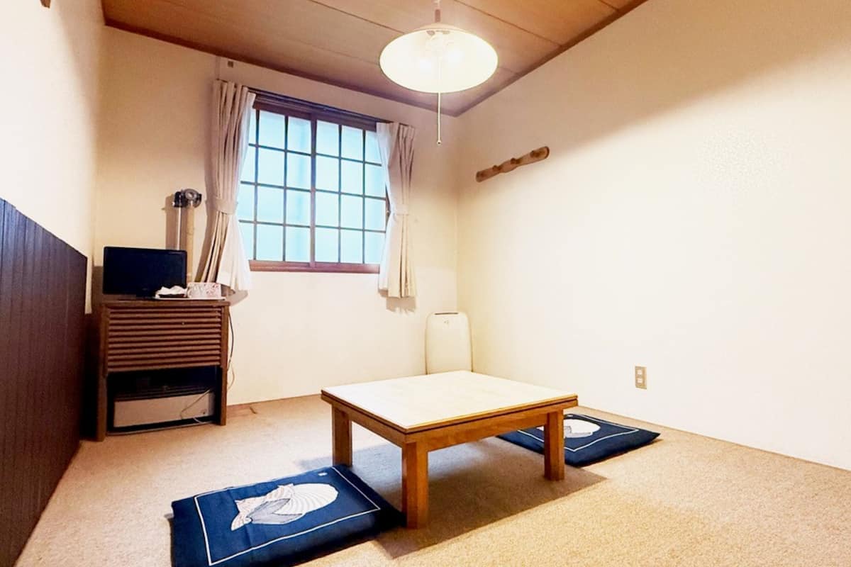 Una habitación con una mesa de madera, una ventana con cortinas, una lámpara y un mueble pequeño.