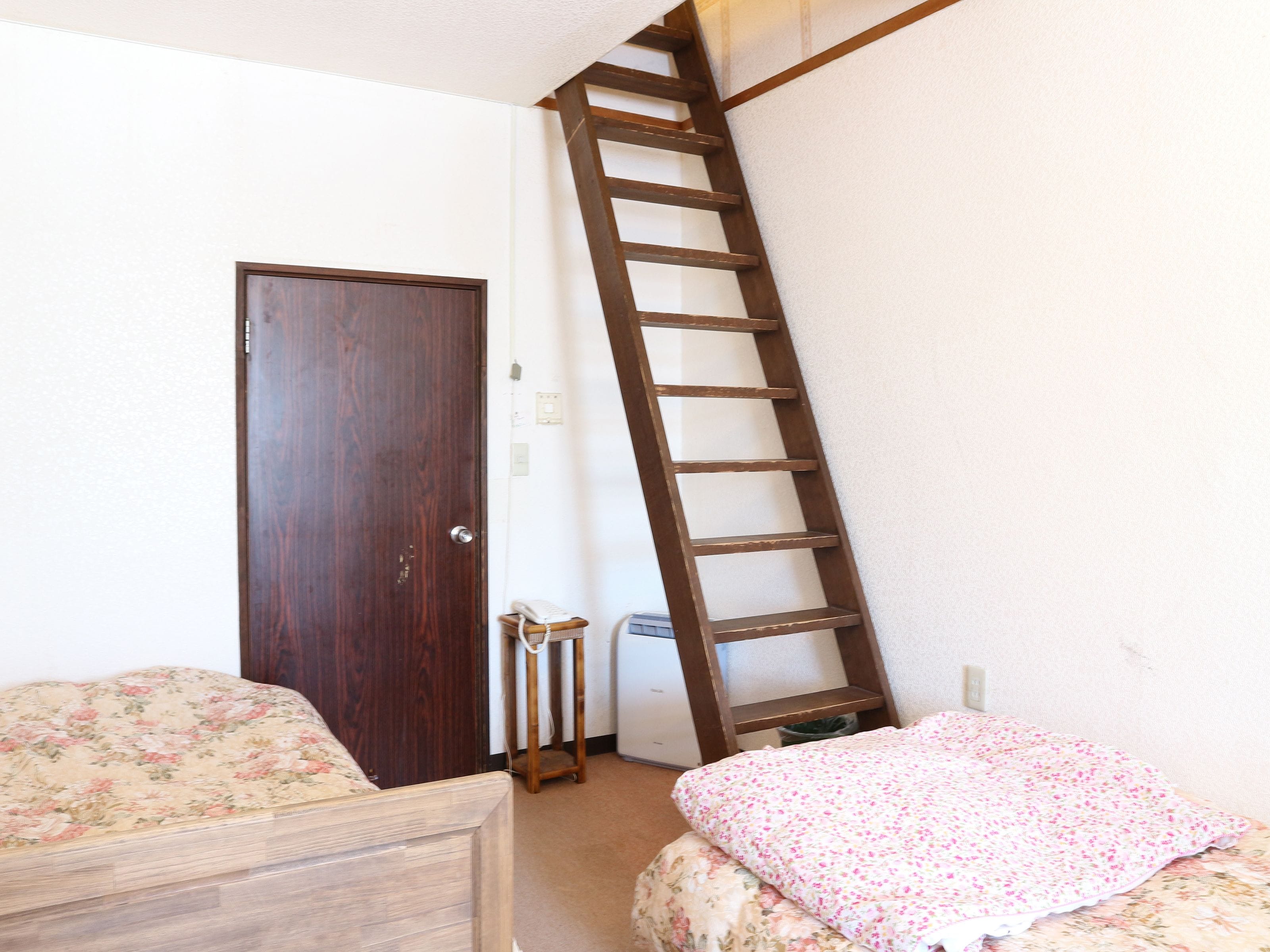 A bedroom with a wooden door, a wooden ladder, a small bedside table, and a bed with floral bedding.