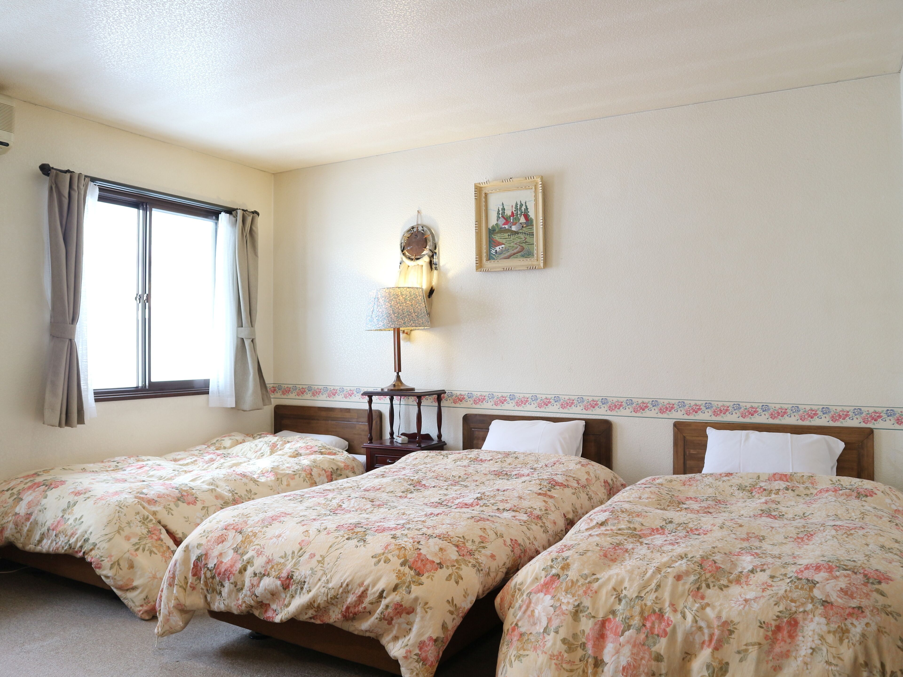 A hotel room with three single beds, a bedside table with a lamp, a window with curtains, and a wall-mounted picture.