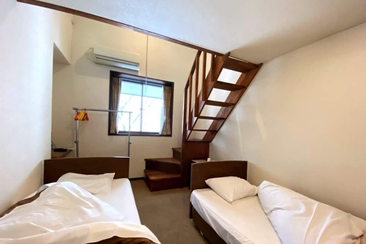 Habitación compacta con dos camas, una escalera de madera, una ventana con cortinas y una pequeña mesa con lámpara.