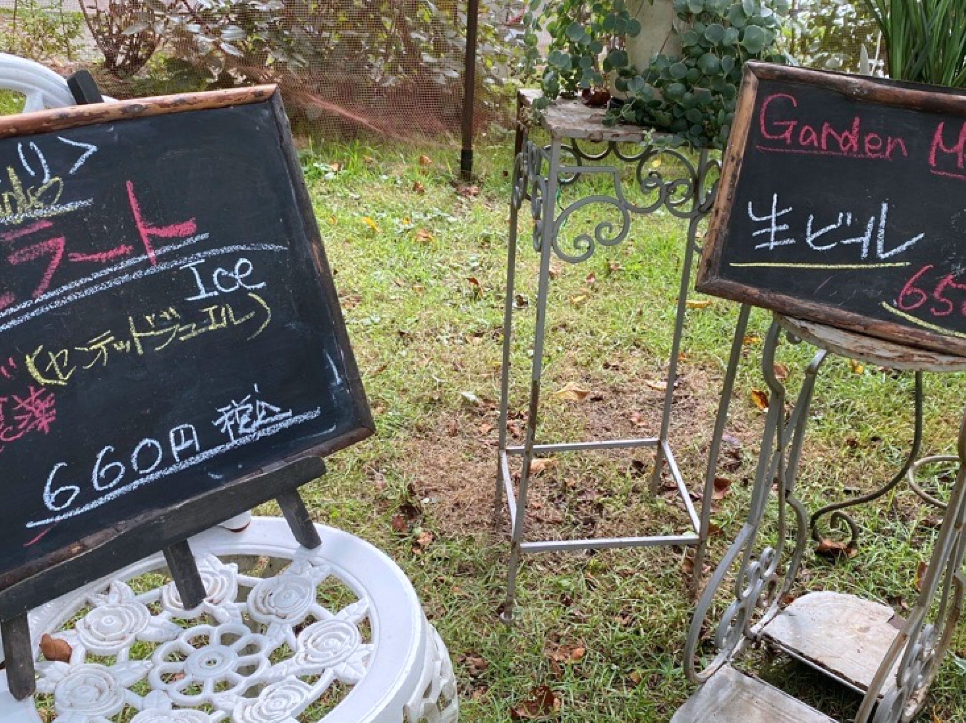 Dos pizarras exteriores con texto, una sobre un soporte blanco y otra sobre un soporte de metal, ubicadas en un jardín con césped y plantas.
