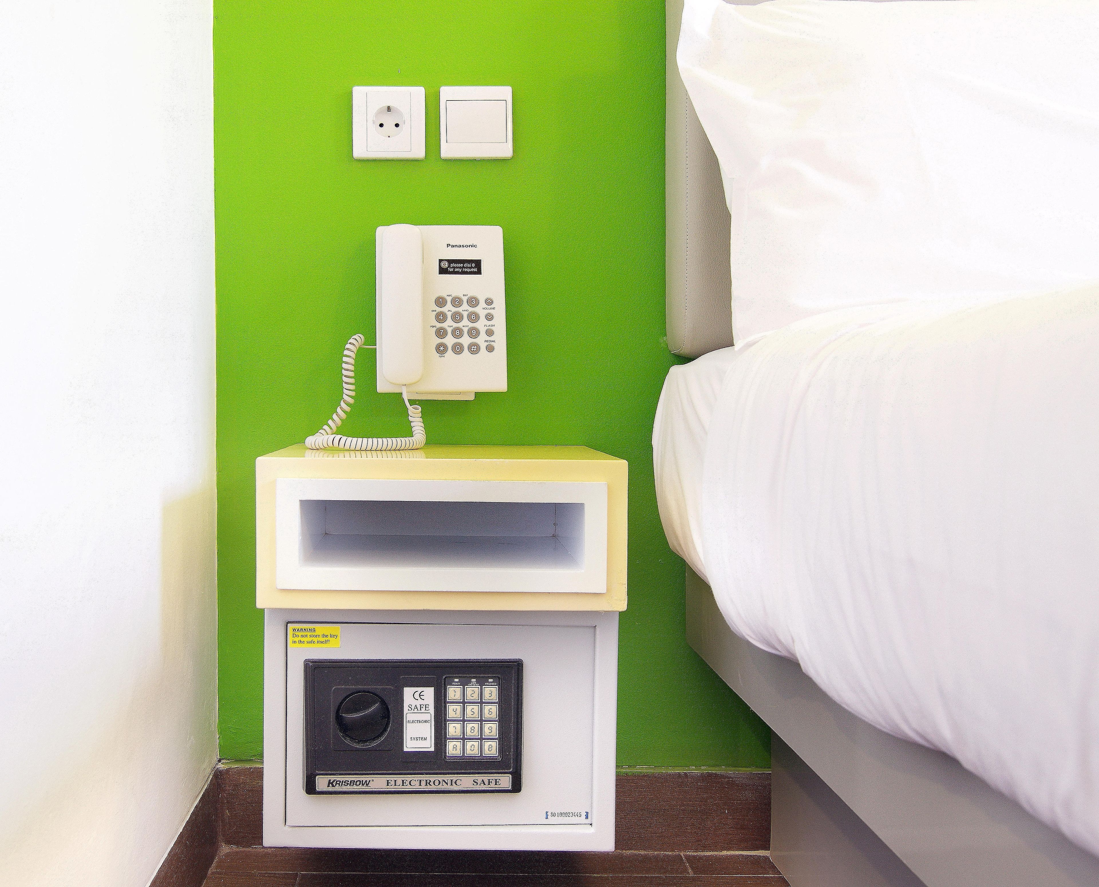A hotel room with a bed, a nightstand with a telephone and a safe, and a green wall.