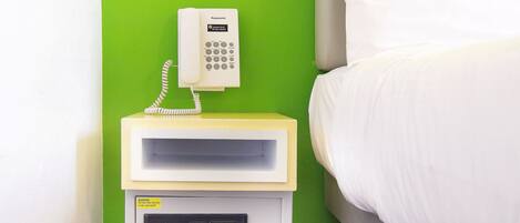 A hotel room with a bed, a nightstand with a telephone and a safe, and a green wall.