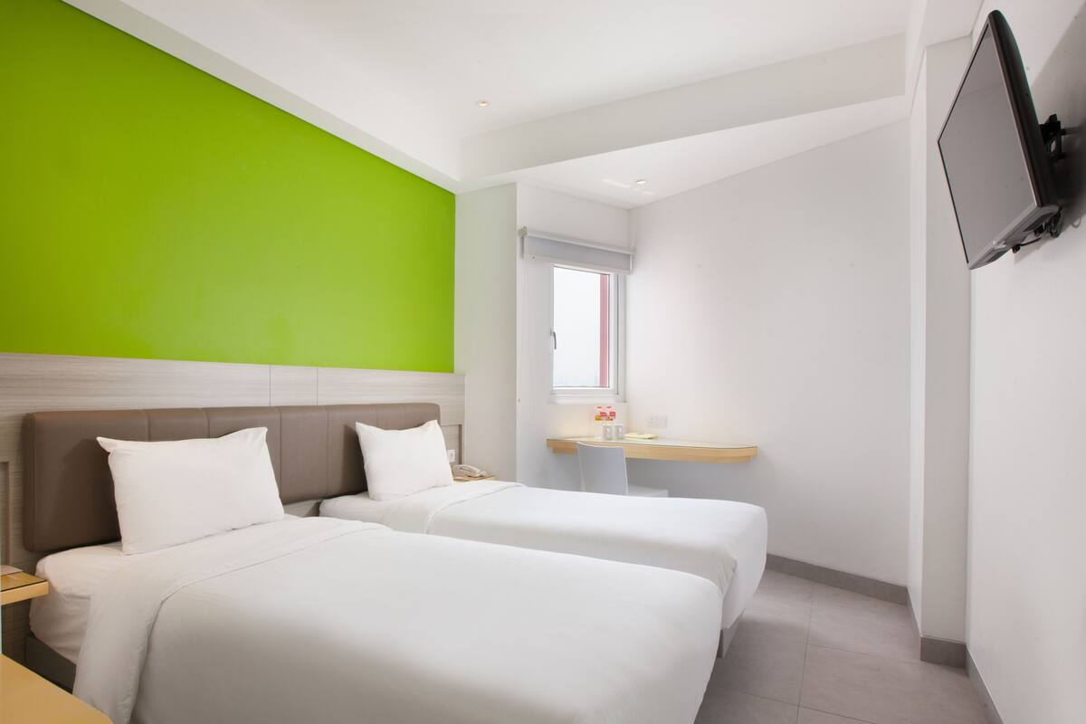 A hotel room with two beds, a desk with a chair, a TV mounted on the wall, and a window providing natural light.