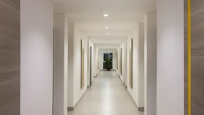 Un pasillo largo y estrecho con paredes blancas y suelo de baldosas, que conduce a una habitación con un ventanal grande y una planta en maceta.