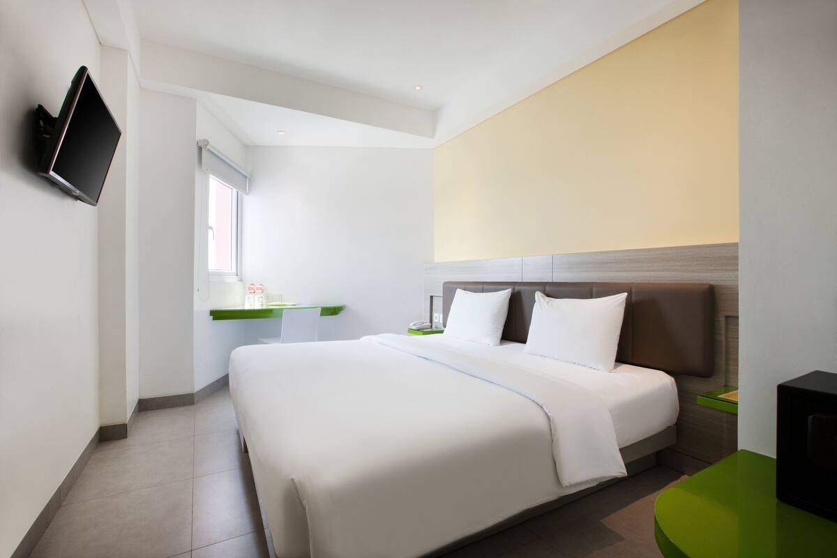 A modern hotel room with a large bed, a flat-screen TV mounted on the wall, a small desk with a chair, and a window providing natural light.