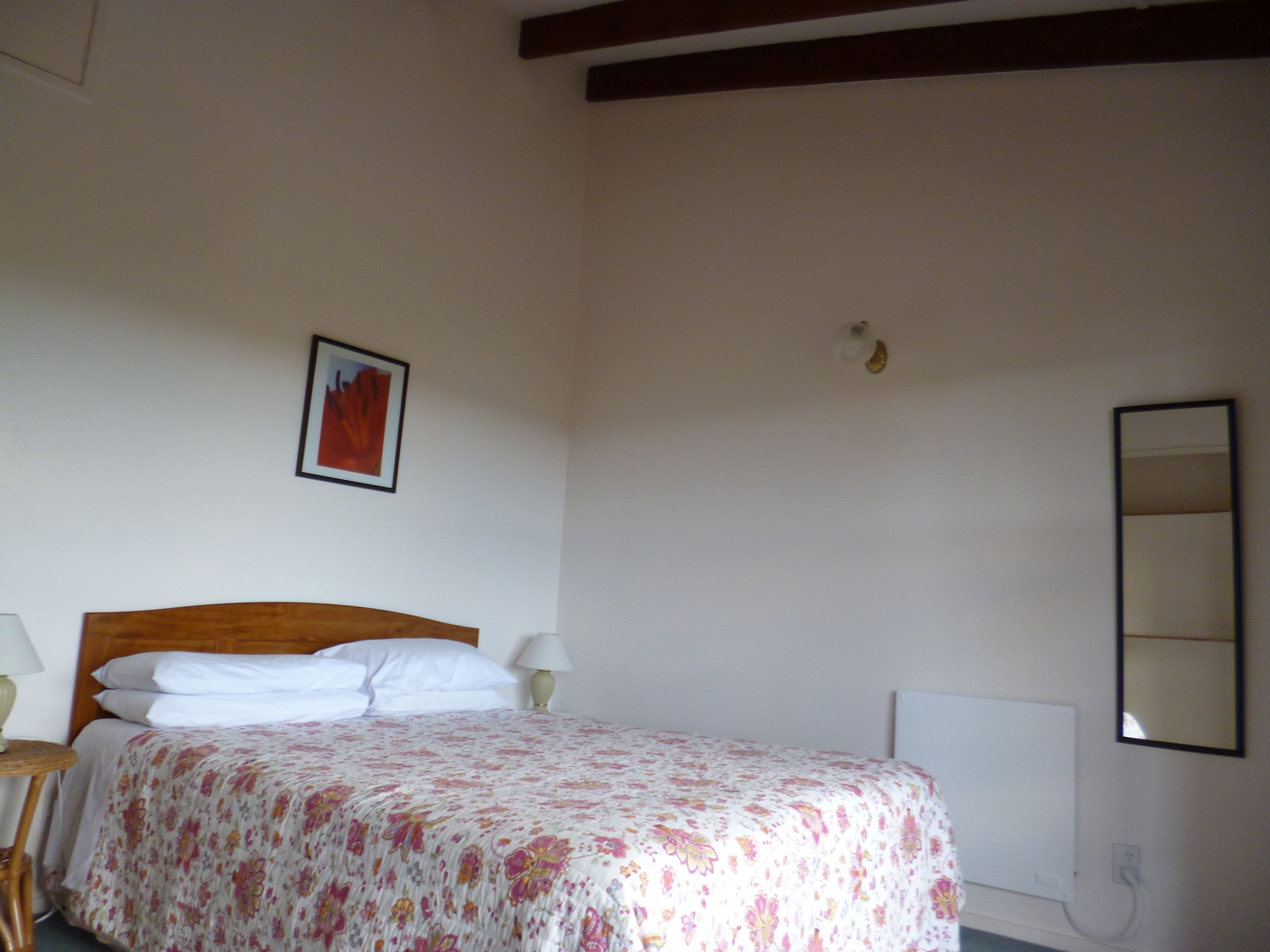 A bedroom with a wooden headboard, a bed with white bedding, a bedside lamp, a wall-mounted mirror, and a framed picture.